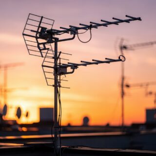Medium shot emphasizing one rooftop broadcast antenna in clear focus with a softfocus cluster of antennas behind it illuminated by the warm glow of sunset.